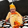 baby, infant, orange_hat, striped_pajamas, pumpkin, smiling, couch, blanket, ghost_pattern, halloween_theme, footed_pajamas, cute, indoor, child, festive, cozy, orange, happy, portrait, holiday