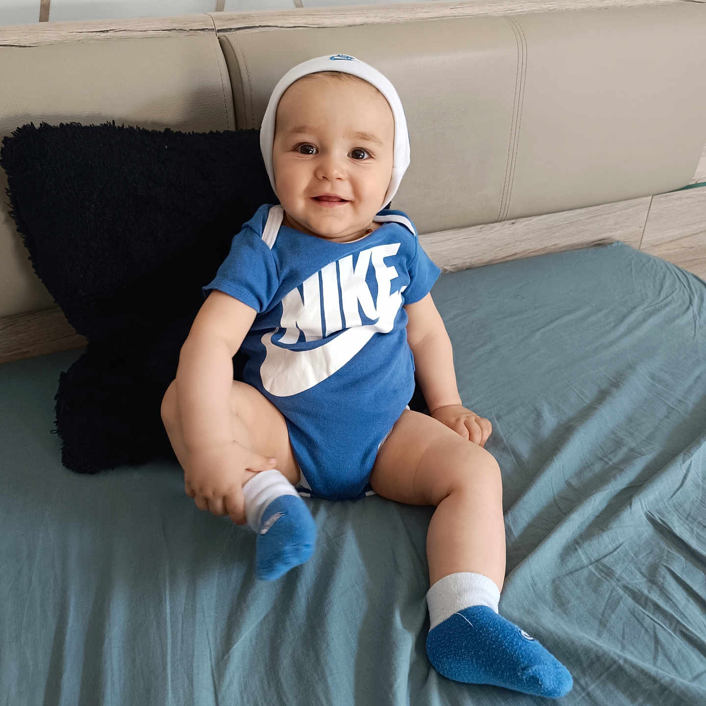 Maël participe au concours pour gagner de l'argent avec cette photo : baby, bed, blue_onesie, casual, child, clothing, comfortable, cute, footwear, happy, headboard, home, indoor, infant, looking_at_camera, person, pillow, sitting, smiling, socks
