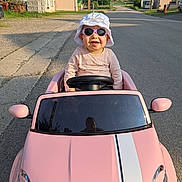 Lise participe au concours pour gagner de l'argent avec cette photo : child, toddler, toy_car, pink_car, sunglasses, hat, smiling, outdoor, street, road, sunny, daytime, vehicle, person, playing, happy, casual_clothing, residential_area, sidewalk, summer