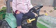 Lise a rejoint le concours — aidez-le/la à gagner de superbes lots ! toddler, child, glasses, pink_sweater, toy_tractor, green, outdoor, concrete_floor, rustic, footwear, pink_sandals, curious, sitting, vehicle, farm, play, young_child, pedal_tractor, casual_clothing, headwear
