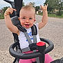 child, toddler, glasses, pink_pants, white_shirt, stroller, outdoor, road, happy, smiling, hands_up, hair, seatbelt, toy_button, nature, trees, grass, bracelet, daylight, person