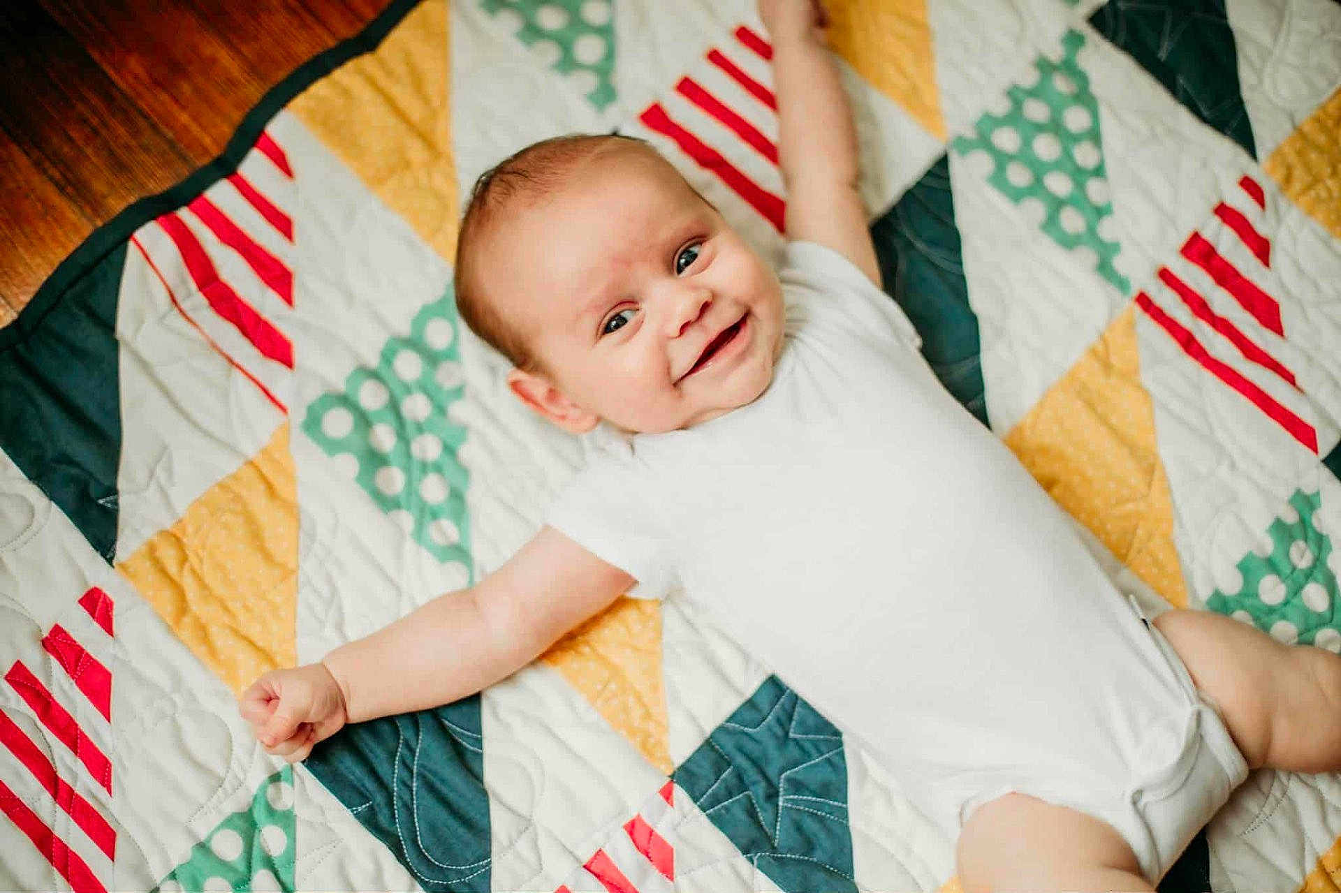 Rowen is registered to the contest to win money with this photo: baby, infant, smile, happy, arms_outstretched, quilt, colorful, pattern, white_onesie, face, child, cute, joyful, skin, lying_down, indoors, textile, fabric, head, portrait