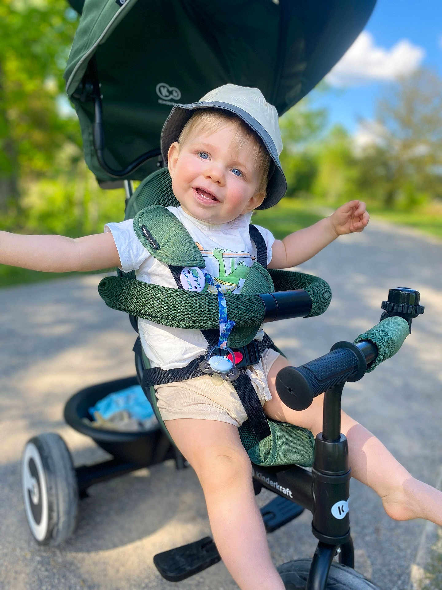 Noah a rejoint le concours — aidez-le/la à gagner de superbes lots ! toddler, child, tricycle, stroller, outdoor, sunny, smiling, bucket_hat, green, blue_sky, trees, gravel_path, playful, happy, summer, shorts, seatbelt, toy, nature, daylight