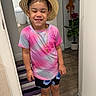 child, smiling, straw_hat, tiedye_shirt, shorts, indoor, happy, person, floor, plant, toy, door, rug, sandals, jewelry, necklace, earrings, wooden_floor, home, casual_clothing
