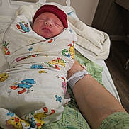 Lorenzo is registered to the contest to win money with this photo: newborn, baby, swaddled, blanket, red_hat, sleeping, hospital_bed, arm, wristband, person, indoor, resting, infant, cute, cozy, medical, patient, care, tender, family