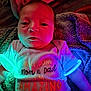 baby, infant, blanket, clothing, face, person, neon_lights, colorful, glow, portrait, cute, text_on_clothing, fabric, soft, newborn, child, lighting, bed, sleepwear, cozy