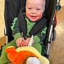 baby, child, smiling, stroller, plush_toy, green_clothing, dinosaur_costume, happy, cute, infant, indoor, person, seat_belt, toy, sock, footwear, blond_hair, blue_eyes, face, portrait