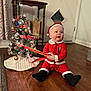 baby, child, santa_claus_outfit, christmas_tree, ornaments, ribbon, holiday, festive, indoor, wooden_floor, curtain, table, door, lights, decoration, red_clothing, black_booties, smiling, celebration, cute