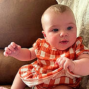 Nora joined the competition — help win amazing prizes! baby, infant, child, cute, orange_dress, checkered_pattern, couch, furniture, indoor, soft_blanket, curious_expression, blue_eyes, chubby_cheeks, short_hair, sitting, hand, skin, portrait, cozy, comfort