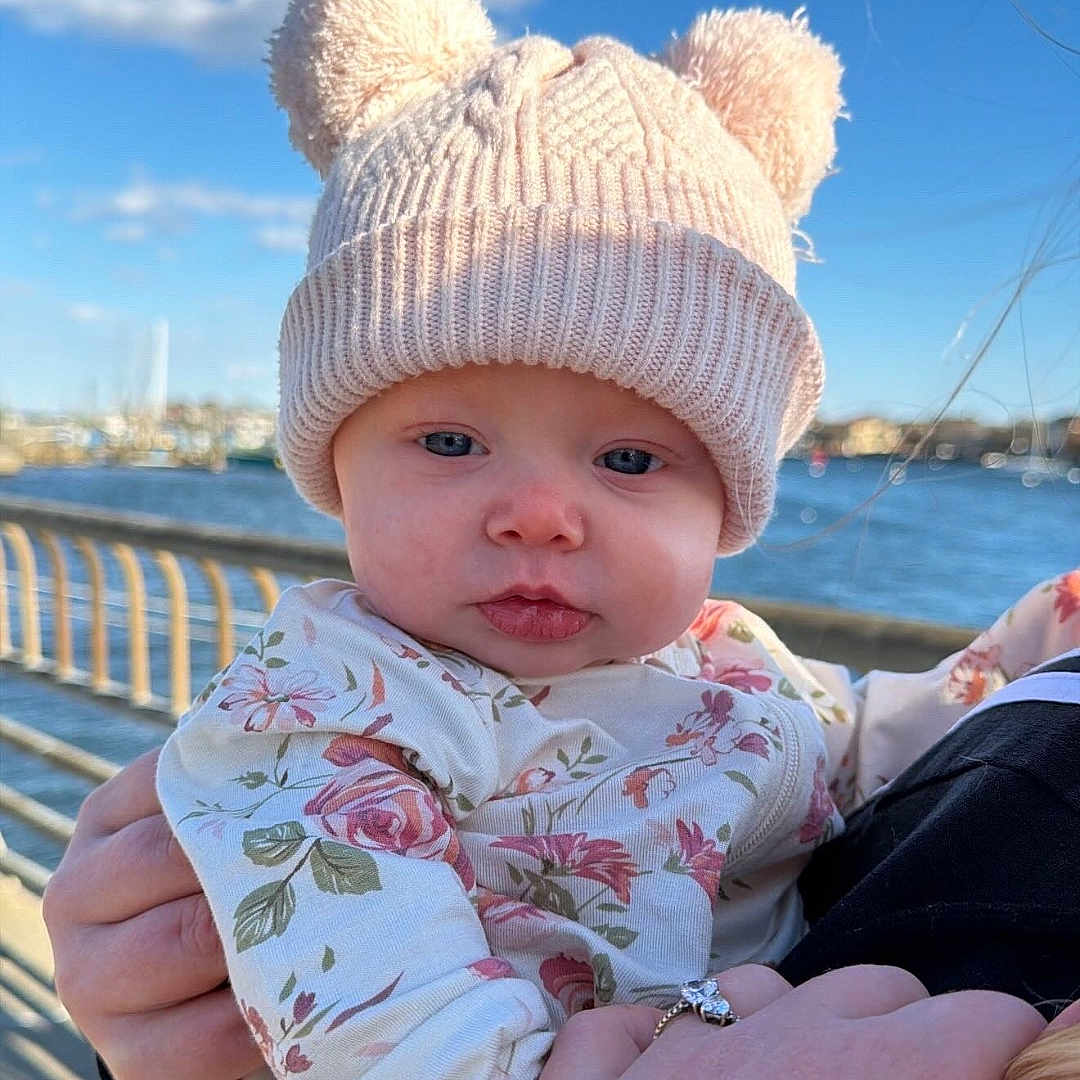 Kiely joined the competition — help win amazing prizes! baby, child, hat, knit_hat, bear_ears, floral_clothing, outdoor, waterfront, sky, clouds, person, holding, hand, portrait, cute, infant, warm_clothing, daylight, blue_sky, nature