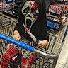 child, shopping_cart, store, mask, scary_mask, toy_knife, adult, man, plaid_pants, socks, aisle, shopping, retail, costume, funny, grocery_store, person, indoor, holding, casual_clothing
