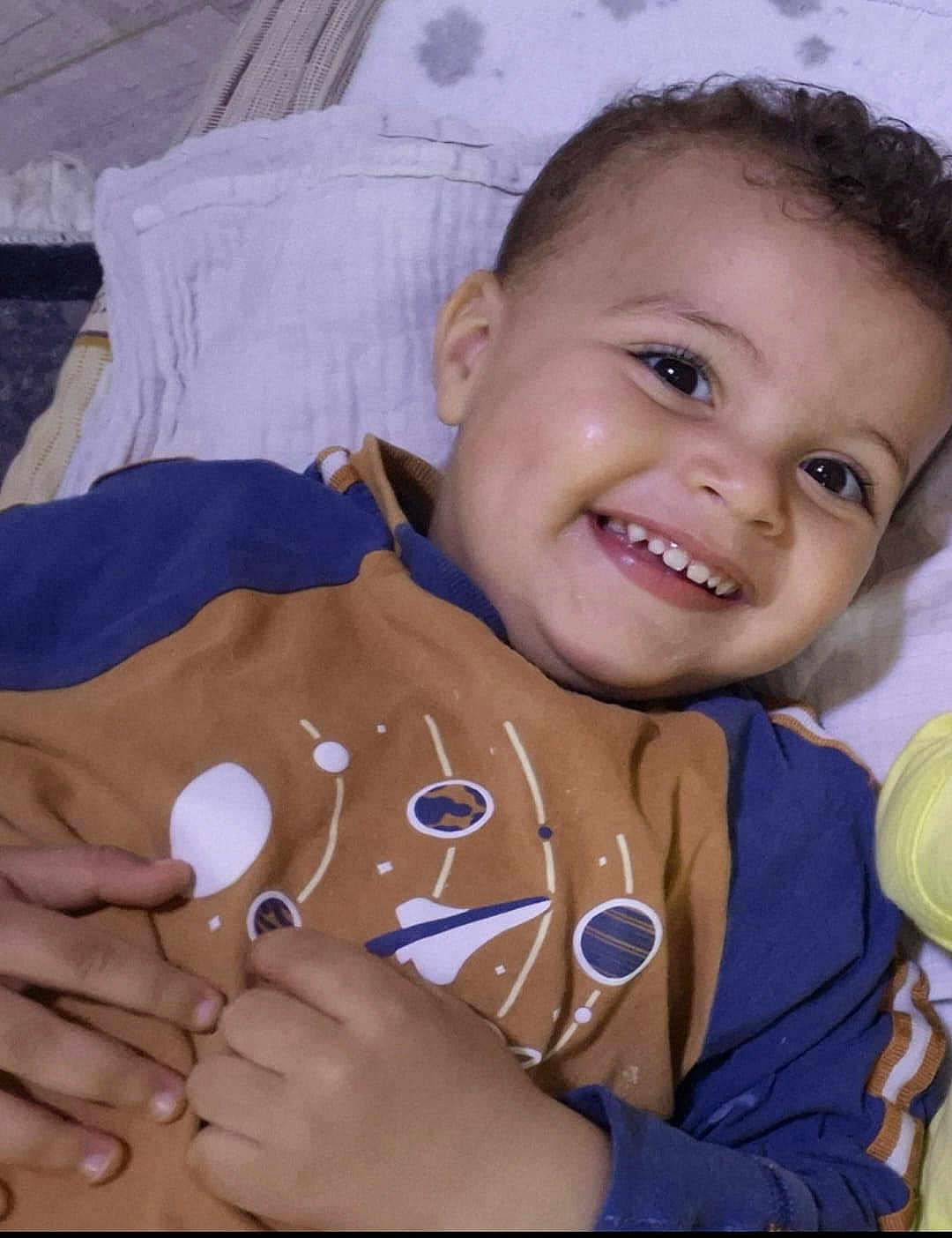 Moumouh participe au concours pour gagner de l'argent avec cette photo : toddler, smiling, child, curly_hair, face, dimples, blue_clothing, brown_clothing, space_design, hand, fingers, lying_down, blanket, happy, portrait, indoors, cute, young_child, bright_eyes, cozy