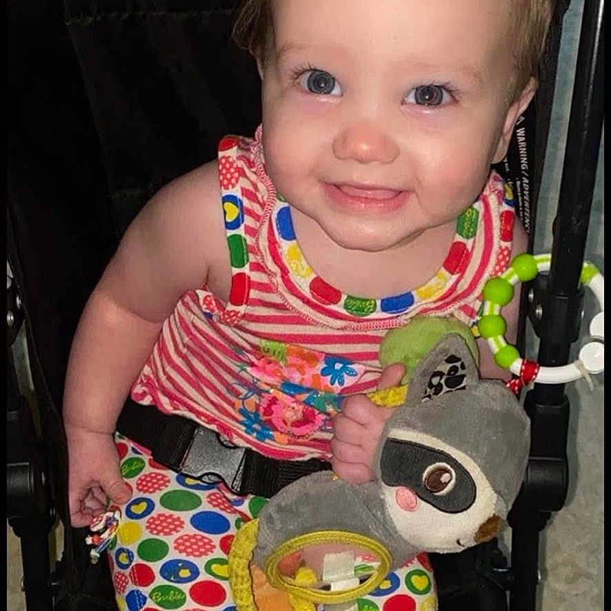 Hayley is registered to the contest to win money with this photo: baby, blue_eyes, child, colorful_clothing, cute, face, hand, happy, indoors, infant, legs, person, plush_toy, polka_dots, red_bow, seatbelt, smiling, striped_shirt, stroller, toy