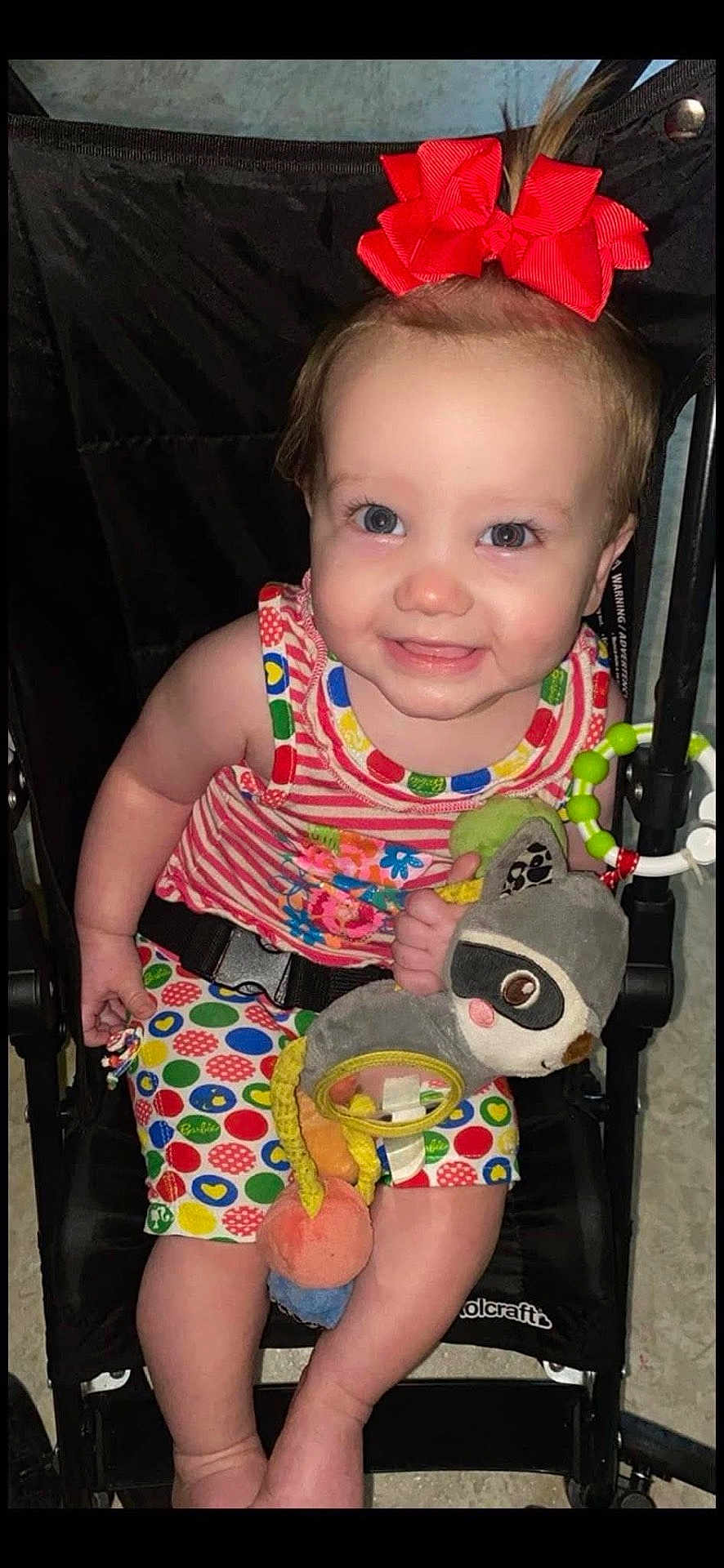 Hayley is registered to the contest to win money with this photo: baby, child, smiling, red_bow, plush_toy, stroller, colorful_clothing, polka_dots, striped_shirt, happy, cute, indoors, person, infant, toy, seatbelt, legs, face, blue_eyes, hand