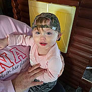 Emilee joined the competition — help win amazing prizes! toddler, child, cap, camo, pink_sweatshirt, adult_hand, indoor, playhouse, wooden_structure, carpet, curious, blue_eyes, long_sleeve, patterned_pants, holding, person, face, smiling, baby, casual_clothing