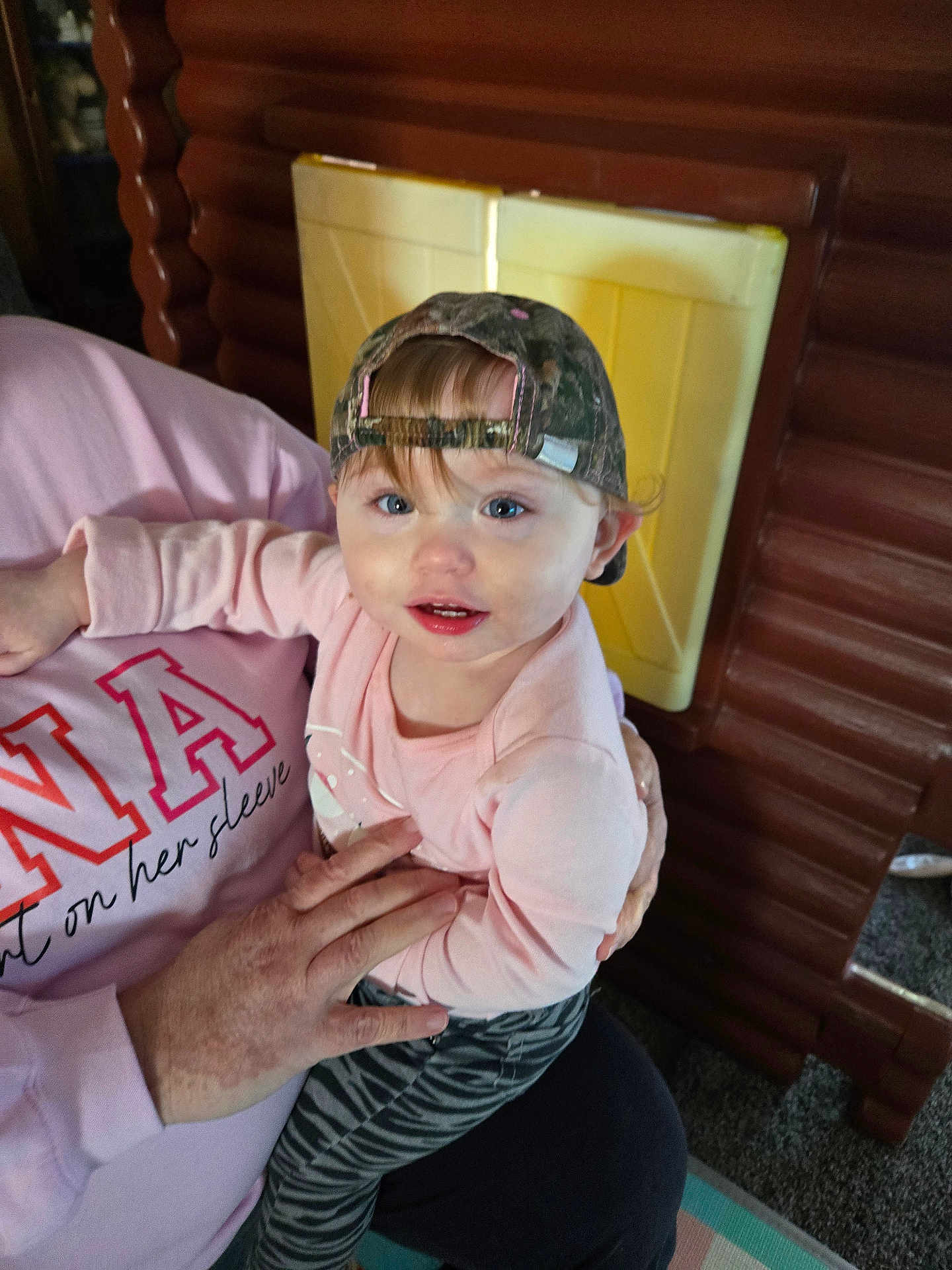 Emilee joined the competition — help win amazing prizes! toddler, child, cap, camo, pink_sweatshirt, adult_hand, indoor, playhouse, wooden_structure, carpet, curious, blue_eyes, long_sleeve, patterned_pants, holding, person, face, smiling, baby, casual_clothing