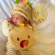 Jad is registered to the contest to win money with this photo: baby, newborn, infant, cute, turkey_costume, hat, plush, blanket, white_bed, sleeping, peaceful, wooden_plaque, birth_details, person, clothing, soft_toys, indoors, baby_outfit, lying_down, portrait