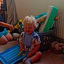 child, toddler, toy, carpet, laundry_basket, window, curly_hair, smile, indoor, sock, plastic_toy, slippers, person, playing, blue_toy, yellow_toy, curtain, radiator, floor, basket