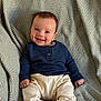 baby, smiling, child, infant, sitting, blue_shirt, cream_pants, blanket, patterned_blanket, happy, cute, indoors, portrait, young_child, small_hands, footwear, cozy, comfortable, face, person