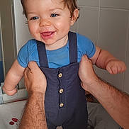 Mylhan a rejoint le concours — aidez-le/la à gagner de superbes lots ! baby, child, smiling, blue_shirt, overalls, hands, indoor, bathroom, tiles, person, cute, happy, standing, skin, hair, face, infant, small_hair_ponytail, arm, adult