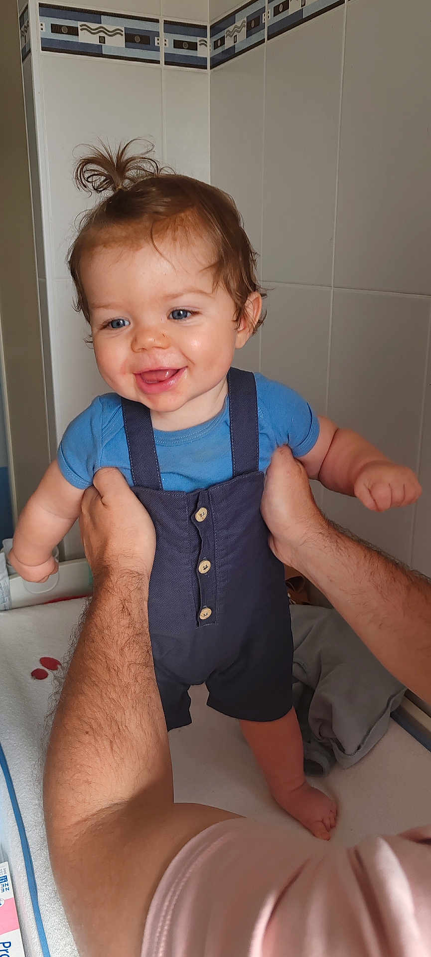 Mylhan a rejoint le concours — aidez-le/la à gagner de superbes lots ! baby, child, smiling, blue_shirt, overalls, hands, indoor, bathroom, tiles, person, cute, happy, standing, skin, hair, face, infant, small_hair_ponytail, arm, adult
