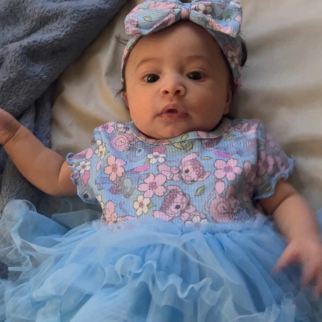 Jayda joined the competition — help win amazing prizes! baby, bed, blanket, blue, child, chubby_cheeks, clothing, curious, cute, dress, face, flower_pattern, headband, indoors, infant, lying_down, person, portrait, soft, tutu