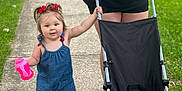 Violet is registered to the contest to win money with this photo: toddler, child, adult, stroller, sidewalk, grass, outdoor, walking, blue_romper, red_bows, sippy_cup, sneakers, person, holding, black_tshirt, tattoos, summer, daylight, cute, playful