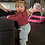 Violet is registered to the contest to win money with this photo: toddler, child, indoor, standing, pigtails, smiling, denim_pants, sneakers, couch, baby_walker, floor, person, hair_tie, casual_clothing, home, portrait, happy, baby, toy, furniture