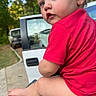 toddler, child, red_shirt, diaper, outdoor, parked_cars, green_surface, curious, portrait, side_view, earrings, nature, trees, daylight, person, baby, short_hair, expression, sitting, side_profile