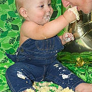 Oliver joined the competition — help win amazing prizes! baby, child, adult, cake, denim_overalls, celebration, messy, feeding, smiling, face, hand, foot, green_background, birthday, food, fun, happy, indoor, person, playful