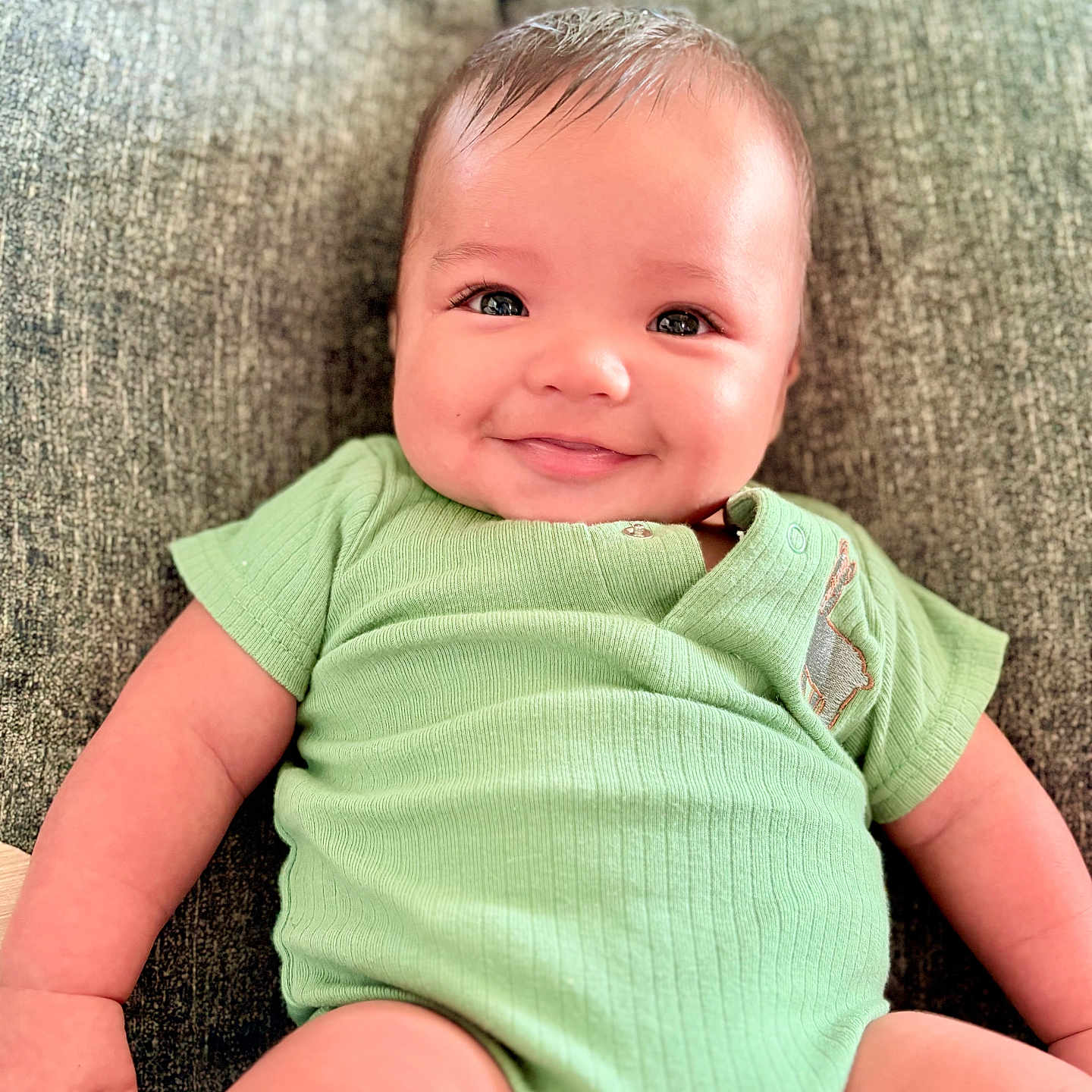 Kyle is registered to the contest to win money with this photo: arm, baby, chubby_cheeks, comfort, couch, cute, face, green_onesie, hand, happy, indoors, infant, leg, person, portrait, relaxation, sitting, skin, smile, young_child