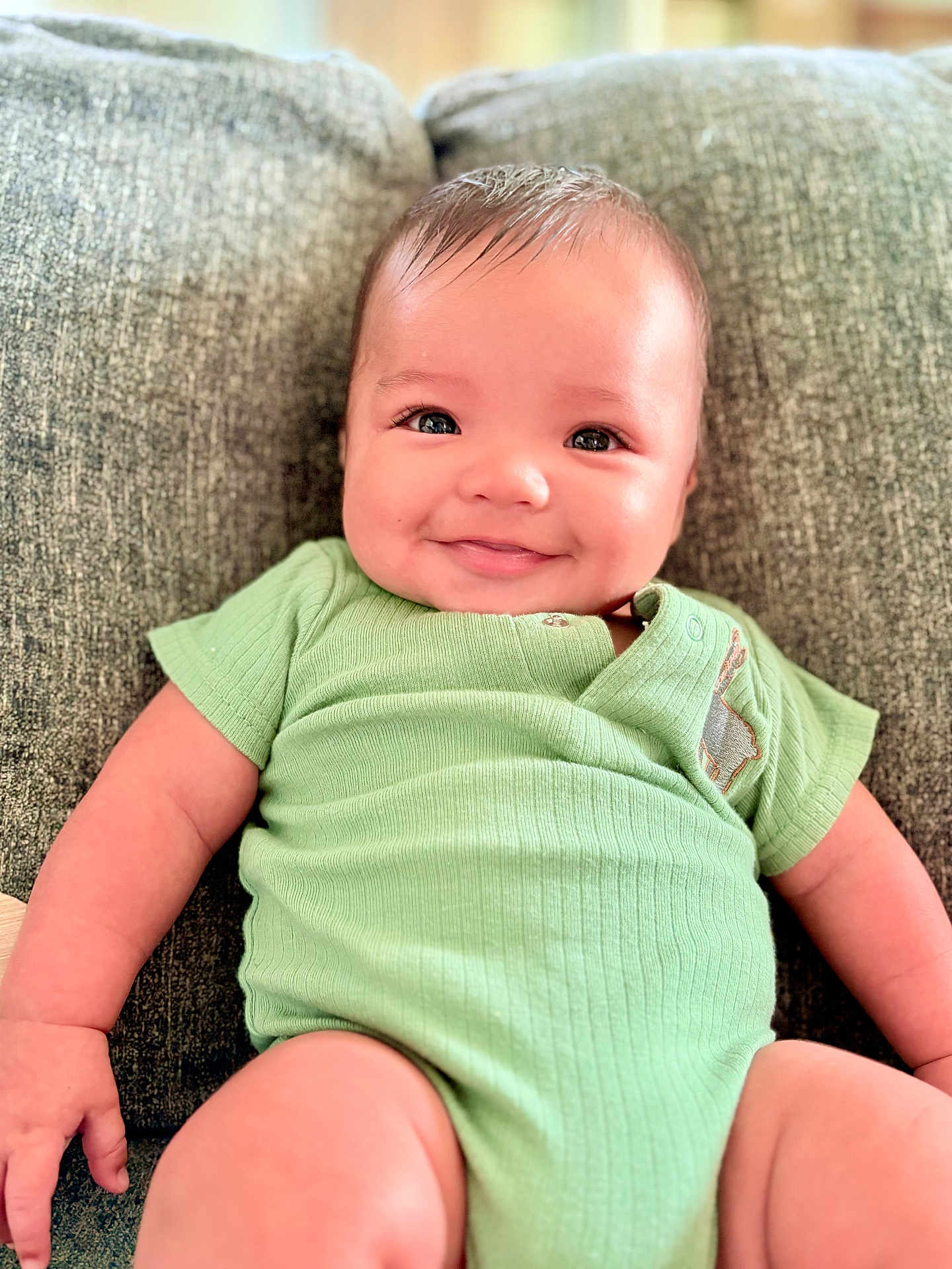 Kyle is registered to the contest to win money with this photo: baby, infant, smile, green_onesie, couch, happy, chubby_cheeks, face, person, sitting, indoors, cute, skin, arm, hand, leg, portrait, young_child, comfort, relaxation