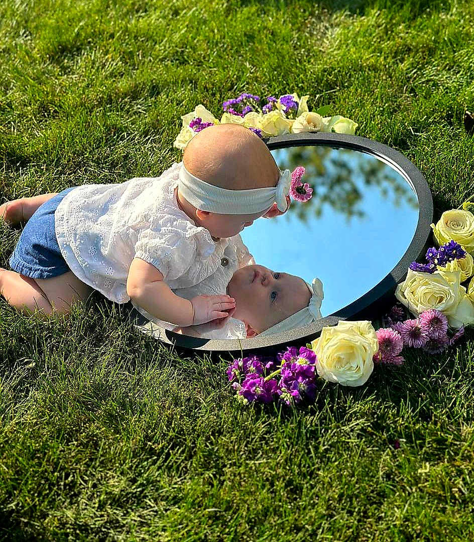 Polina is registered to the contest to win money with this photo: baby, child, reflection, mirror, grass, flowers, headband, outdoor, sunlight, white_blouse, purple_flowers, white_flowers, nature, portrait, curious, crawling, grass_field, closeup, baby_girl, spring