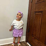 Merlyn is registered to the contest to win money with this photo: child, girl, smile, purple_skirt, headband, white_shirt, sandals, wooden_door, hardwood_floor, wall, baseboard, standing, happy, indoors, clothing, footwear, person, cute, portrait, fashion