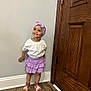 child, girl, smile, purple_skirt, headband, white_shirt, sandals, wooden_door, hardwood_floor, wall, baseboard, standing, happy, indoors, clothing, footwear, person, cute, portrait, fashion