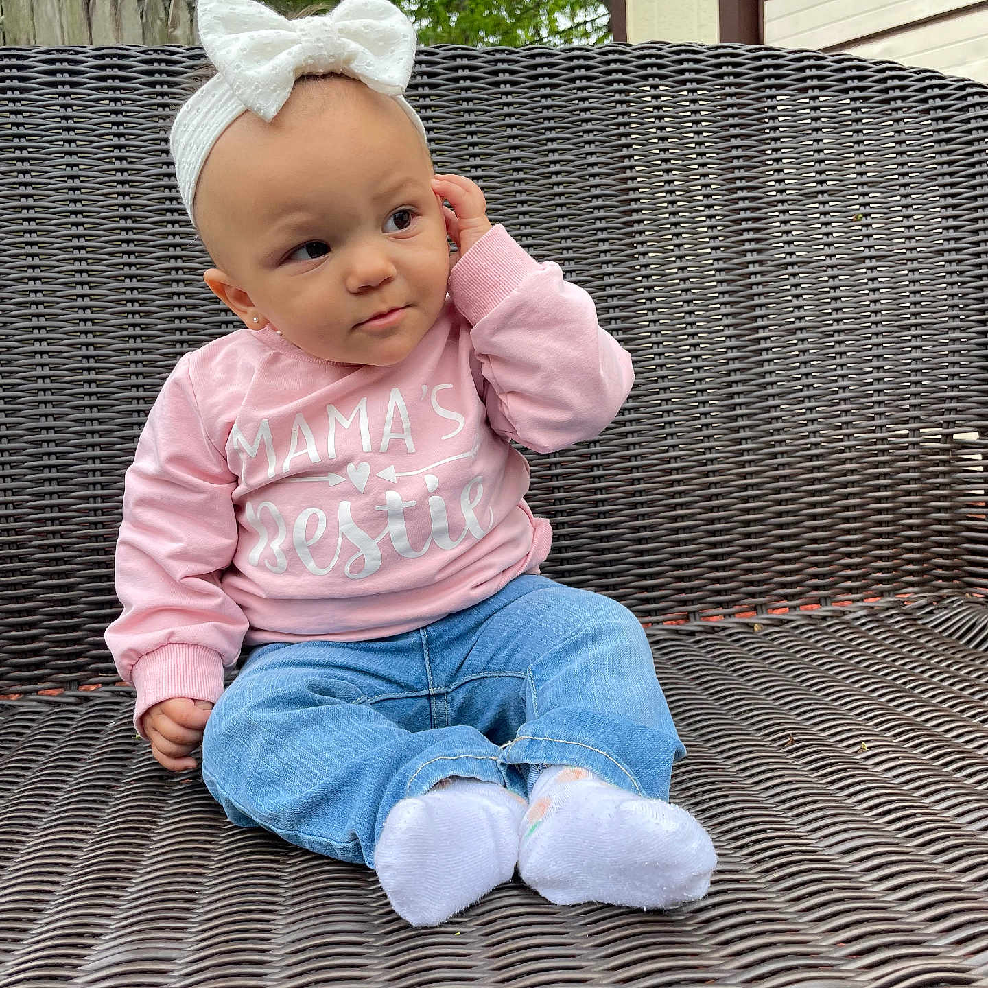 Jennica is registered to the contest to win money with this photo: baby, bow, casual_clothing, child, cute, expression, fence, greenery, headband, jeans, outdoor, person, pink_sweatshirt, portrait, sitting, text_on_clothing, tree, white_socks, wicker_chair, young