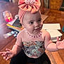 baby, child, pink_bow, headwrap, necklace, table, wooden_table, arms, sitting, indoors, jewelry, cute, smiling, face, person, clothing, black_pants, home, blurred_background, candle