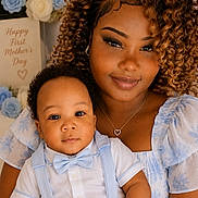 Leo is registered to the contest to win money with this photo: woman, baby, portrait, smile, curly_hair, bow_tie, suspenders, mother, child, flower_arrangement, blue, white, celebration, happy, love, family, indoor, clothing, necklace, skin