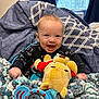 baby, smiling, plush_toy, blanket, window, curtains, indoor, child, happy, knitted_blanket, toy, person, face, blue, red, yellow, furniture, light, cozy, cute