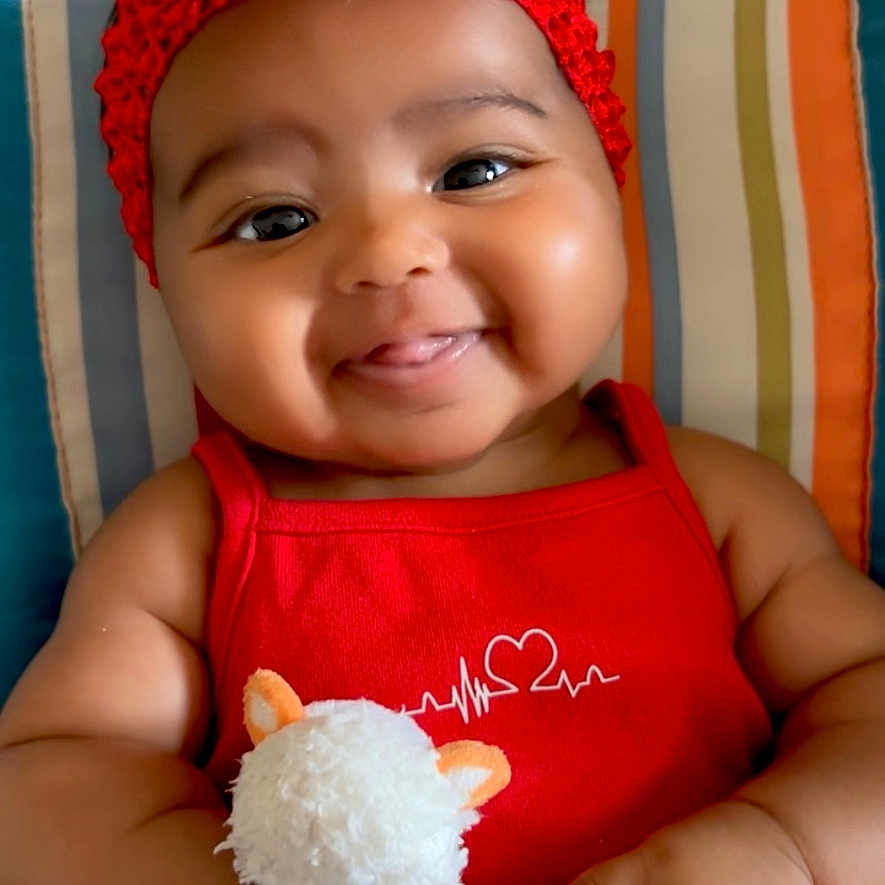 Dayana participe au concours pour gagner de l'argent avec cette photo : baby, child, chubby_cheeks, closeup, colorful_background, cute, face, flower, hand, happy, indoors, infant, person, plush_toy, portrait, red_headband, red_outfit, smiling, striped_cushion, toy