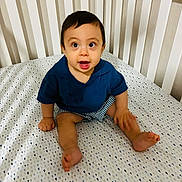 Ricardo is registered to the contest to win money with this photo: baby, crib, child, infant, blue_shirt, striped_shorts, patterned_sheets, white_crib, sitting, happy, cute, indoors, person, smiling, young_child, face, foot, hand, room, furniture