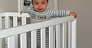 Ricardo joined the competition — help win amazing prizes! baby, crib, child, infant, smiling, striped_clothing, onesie, indoor, door, white_crib, carpet, standing, person, young_child, baby_boy, happy, grip, room, neutral_wall, home