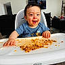 toddler, high_chair, messy, food, smiling, happy, indoor, child, face, clothing, table, tray, furniture, living_room, person, expression, meal, hair, seat, playful