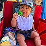 Ricardo joined the competition — help win amazing prizes! baby, child, stroller, red, hat, bib, teething_toy, toy, blanket, outdoor, sunlight, dinosaur_print, person, hand, clothing, happy, smile, seatbelt, fabric, summer