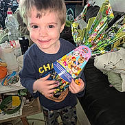 Matëo a rejoint le concours — aidez-le/la à gagner de superbes lots ! child, smiling, indoor, gift, colorful_wrapping, cartoon_characters, barefoot, table, couch, clutter, toy, pajamas, face, person, young_boy, happy, living_room, plastic_bottle, cup, furniture