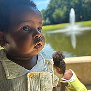 Elian joined the competition — help win amazing prizes! toddler, child, portrait, outdoor, pond, fountain, greenery, sunny, daylight, curly_hair, shirt, yellow_dress, nature, water, reflection, person, background_blur, closeup, expression, summer