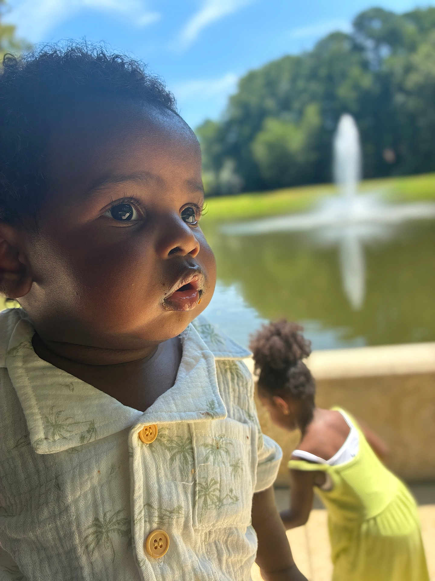 Elian joined the competition — help win amazing prizes! toddler, child, portrait, outdoor, pond, fountain, greenery, sunny, daylight, curly_hair, shirt, yellow_dress, nature, water, reflection, person, background_blur, closeup, expression, summer