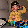 toddler, child, toy_car, cap, yellow_clothing, couch, gray_furniture, indoor, smiling, playing, person, face, hand, seat, home, cute, happy, car_toy, casual, young_child