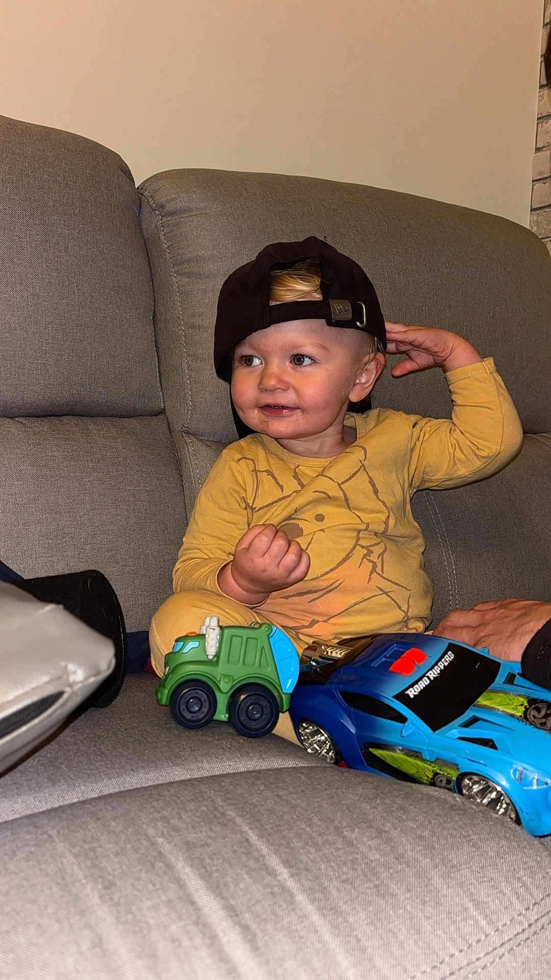 Liam a rejoint le concours — aidez-le/la à gagner de superbes lots ! toddler, child, toy_car, cap, yellow_clothing, couch, gray_furniture, indoor, smiling, playing, person, face, hand, seat, home, cute, happy, car_toy, casual, young_child