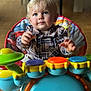 Liam participe au concours pour gagner de l'argent avec cette photo : child, toddler, blonde_hair, blue_eyes, plaid_shirt, baby_toys, colorful, indoor, wooden_floor, high_chair, play, smiling, cute, person, face, hand, seat, furniture, young_child, happy